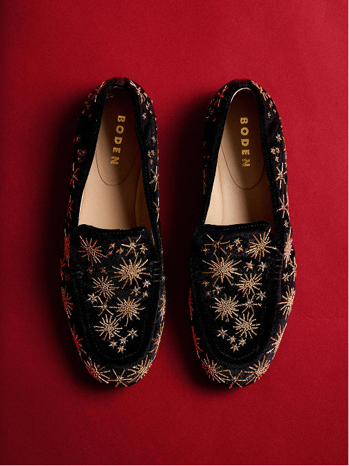 Ein Paar BODEN-Slipper aus schwarzem Samt mit goldgestickten Sternenmustern, flach vor einem einfarbigen roten Hintergrund präsentiert.